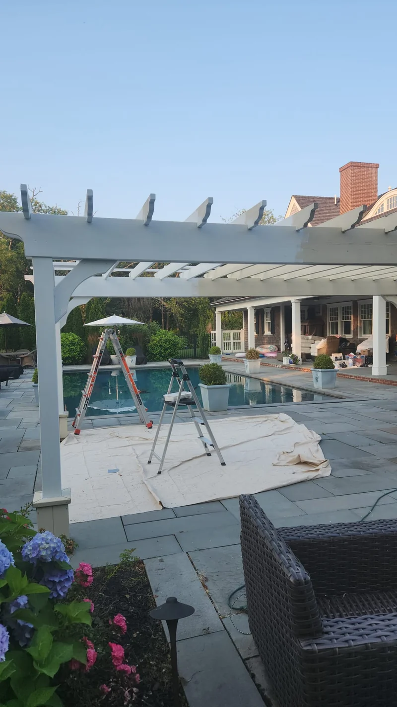 White pergola above a stone pool terrace, with ladders and a protective drop cloth during exterior work