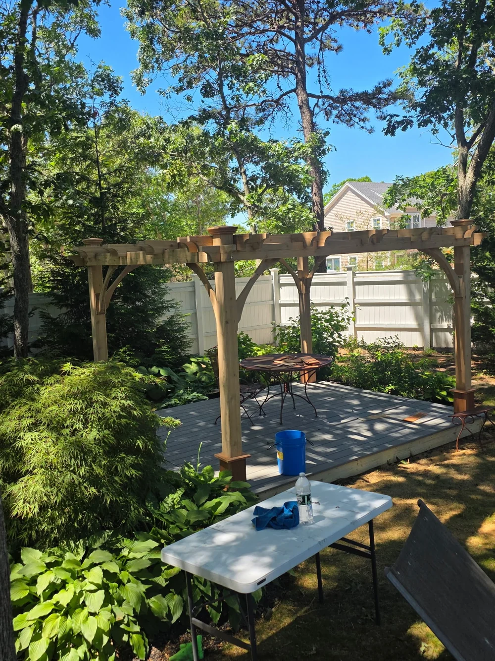 After: refreshed wood pergola with capped posts above a gray deck in a leafy garden, with work materials still present