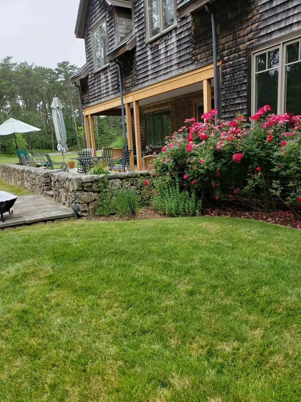 Shingle-clad home with light wood porch posts and horizontal trim beside a stone terrace and flowering garden